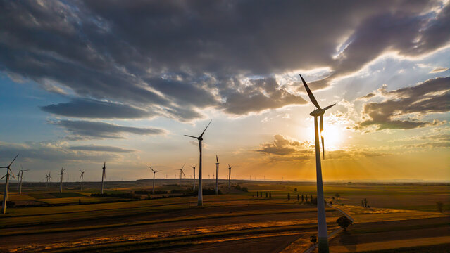 Wind turbine beside sunbeams over shaded farmland. Gray clouds frame the sun behind a foreground tower, with parallel crop strips and a winding track below.