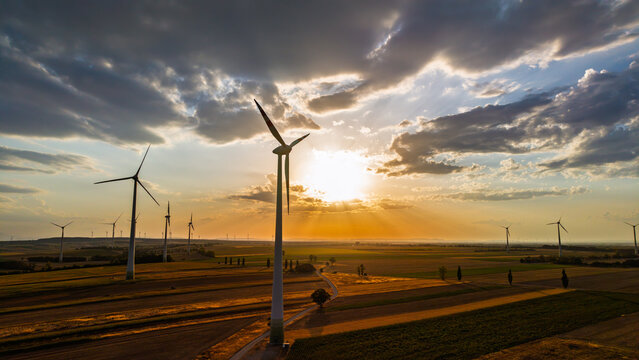 Wind turbine with the sun behind its rotor. Gray clouds frame the light above a white tower, parallel fields and long diagonal shadows.