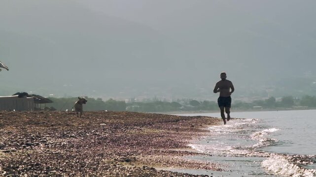 A slow motion video of an eight year old female one eyed pitbull running together with her athletic forty year old male owner on a sunny morning sandy beach.