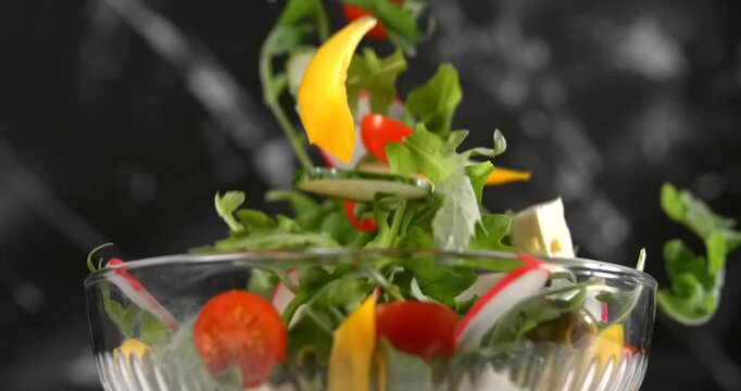 Fresh vegetable salad ingredients falling into glass bowl in slow motion on black background