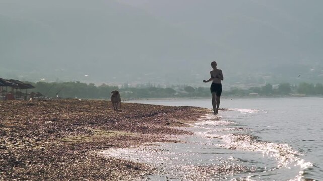 A slow motion video of an eight year old female one eyed pitbull running together with her thirty year old female owner on a sunny morning sandy beach.