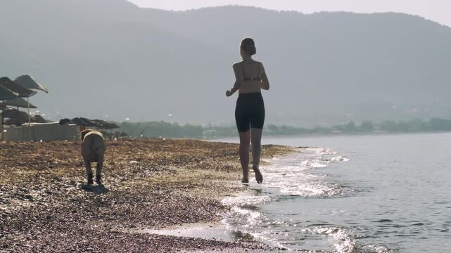A slow motion video of an eight year old female one eyed pitbull running together with her thirty year old female owner on a sunny morning sandy beach.