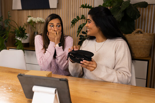 Diverse female friends sitting at wooden table in cozy room, holding open black case beside tablet