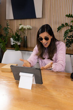 Indian woman wearing shades and pink top reacting with surprise toward tablet stand in workspace