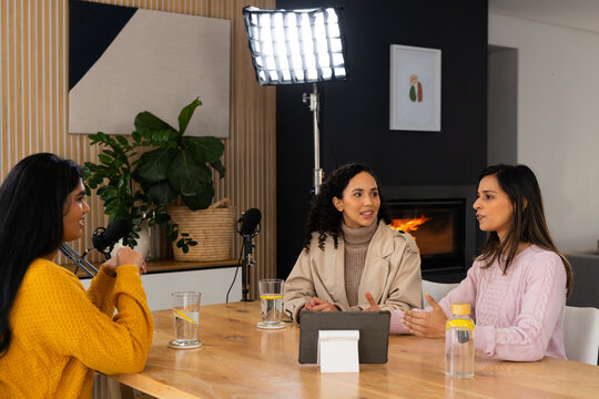 Diverse female colleagues recording in home studio wearing knit sweaters with mics, tablet