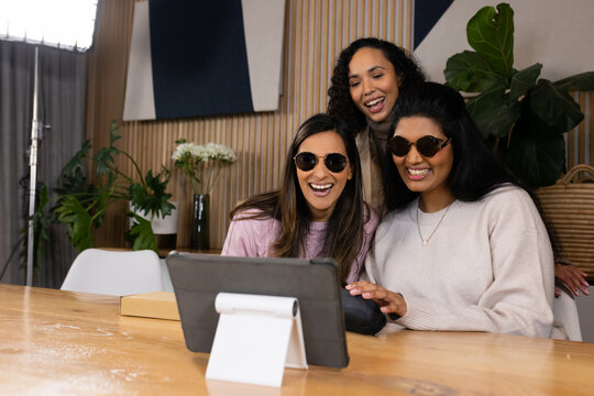 Diverse female friends leaning over tablet on wooden table in studio, smiling and using mouse