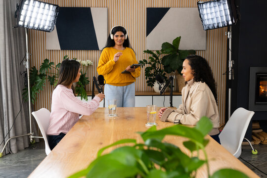 Diverse female colleagues recording discussion at long table with mics tablet yellow sweater host