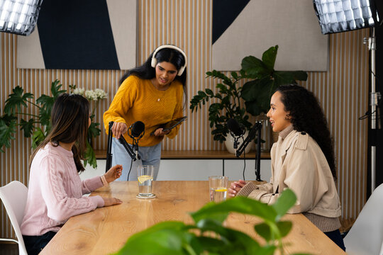 Diverse female podcast team recording at studio wooden table, adjusting microphone and using tablet