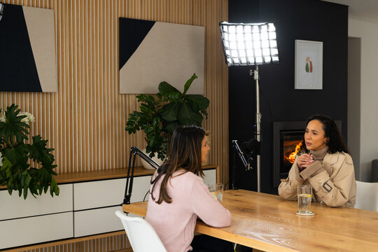 Diverse female duo talking in studio at wooden table with boom mics, pink sweater, trench coat