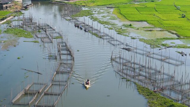 Aerial view of Lake Rawa Pening featuring fish farming nets and a boat navigating the water surrounded by lush green rice paddies in Waduk Rawapening, Central Java, Indonesia.