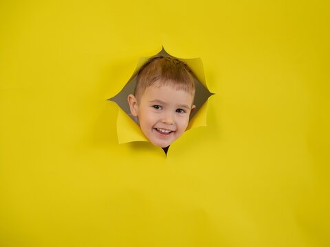 A mischievous boy peeks out through a hole in a yellow cardboard background. 