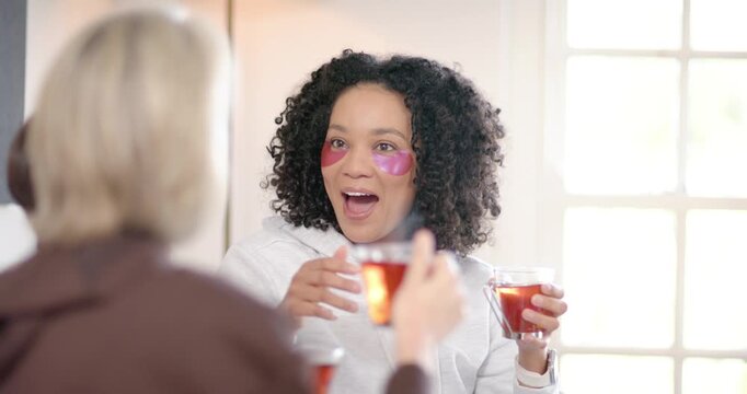 African woman in hoodie adjusting pink eye patches toasting cups with blonde friend at kitchen