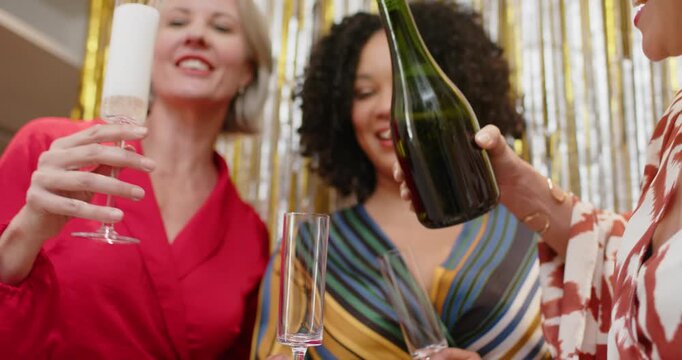 Diverse women smiling, woman on right pouring green bottle, filling flute at gold tinsel for toast