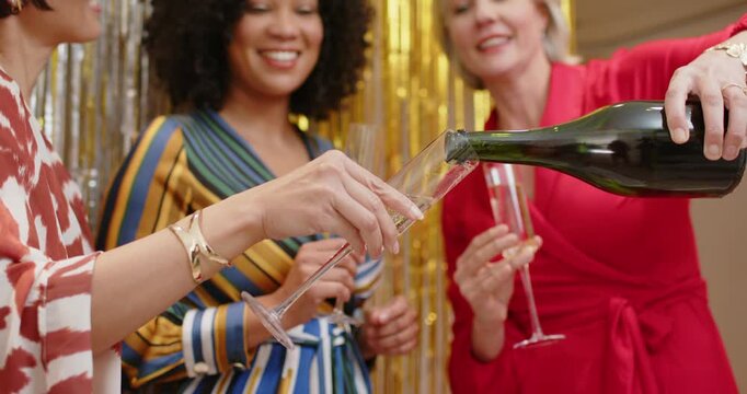 Three women in dresses pouring champagne after bottle lift filling flutes for party at gold fringe