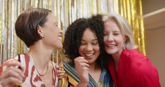 Diverse women friends leaning in at party, hugging and blowing gold horns at fringe backdrop