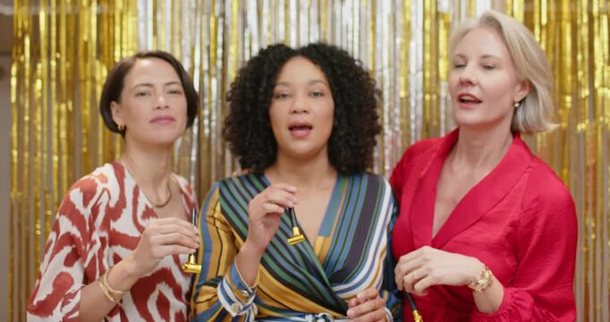 Diverse female friends center smiling raising and blowing gold blowers in gold tinsel celebrating