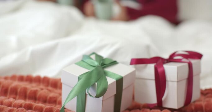 Diverse couple noticing gift boxes and laughing while sipping mugs on bed at christmas