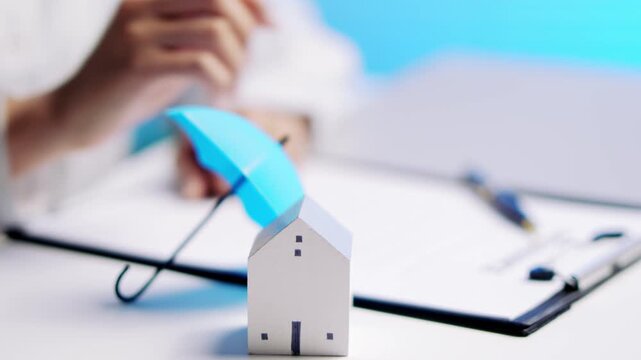 Professional house insurance coverage concept, hand holding small umbrella over model building protection plan, finance risk safety document writing