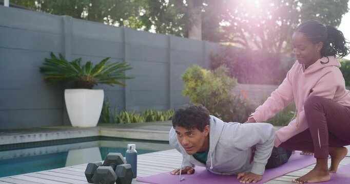 African American workout partners man doing push-ups on purple mat poolside, woman steadying torso