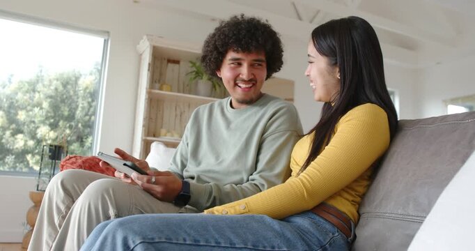 Diverse couple on sofa in living room, man tapping tablet with remote sharing screen