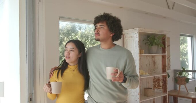 Diverse couple standing by glass door spotting movement outside, both holding mugs, woman pointing