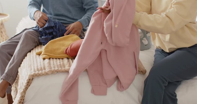 Placing gray laundry basket on bed, African American couple folding hoodie and plaid pants to sort