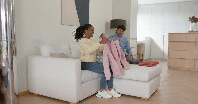African American couple reacting to phone, folding pink sweater while sorting laundry on white sofa