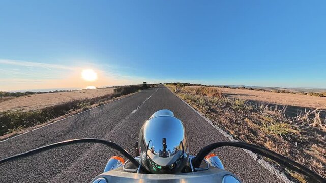 Motorcycle riding along a straight rural road at sunset, surrounded by open countryside and warm golden evening light, creating a scenic road travel atmosphere.
