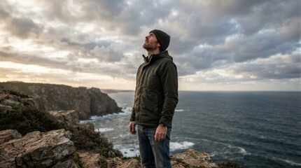 Fototapeta premium Man stands on cliff's edge, gazing at ocean. Clouds swirl above as sun breaks through. Windy, rugged coast behind him. He looks up, relaxed, thoughtful. Perfect for travel, adventure, or nature themes