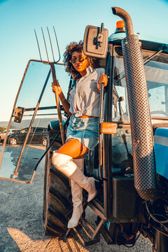 Confident woman stands on farm tractor steps holding pitchfork under bright golden sunlight, creating bold rural lifestyle portrait with industrial machinery, open sky, and outdoor parking area.