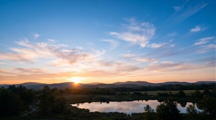 Fototapeta premium Golden sun dips low over distant hills. Sky blushes with soft orange and blue hues. Clouds drift gently across the evening horizon. Still lake mirrors the sun's fading glow. Trees frame the scene