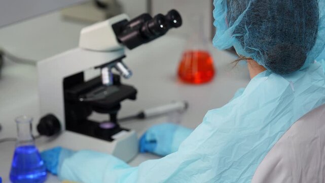 Over-the-shoulder shot of a scientist in a blue hair net and protective gown adjusting microscope objectives and focus, with flasks of red and blue liquid on the lab bench