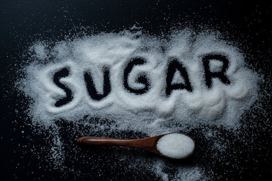 Word sugar written in white granulated sugar on black background. Conceptual top view of word SUGAR written on white spilled granulated sugar with wooden spoon on dark black textured surface