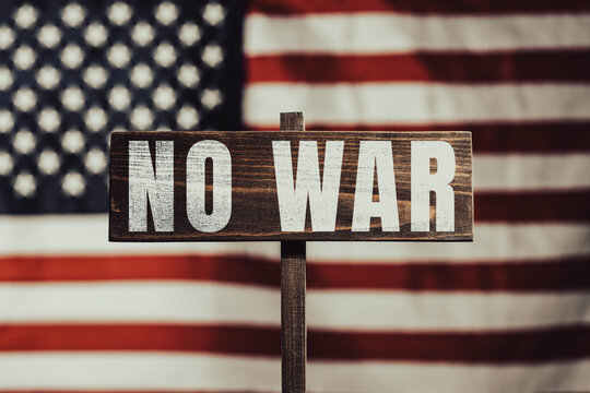 No war protest sign with American flag background. Conceptual photo of wooden sign with text NO WAR closeup against blurred United States flag background