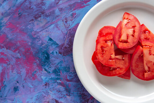 Fresh tomato slices rest on a white plate. Vibrant red hues contrast with textured purple-blue background. Simple, clean composition draws focus to natural ingredients. Ideal for health, cooking