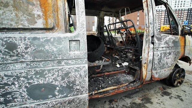 Charred minibus displays heavy damage from destructive fire with peeling paint and rust. Gutted automobile stands parked outside showing exposed frame alongside ruined passenger compartment.