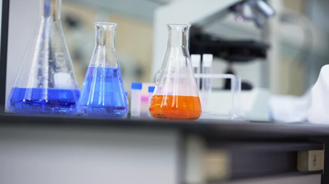 Close-up of chemistry flasks filled with blue and orange liquid on a laboratory bench as a blue gloved hand removes a test tube from the rack and then leaves the frame