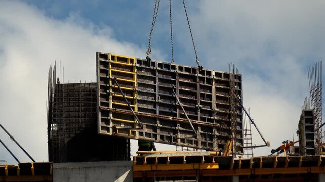 Crane lifts concrete formwork panel above unfinished structure while worker guides heavy steel component against cloudy blue sky. Shuttering panel hovers carefully over building framework.