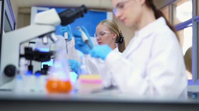 Two female scientists working side by side at microscopes, a blonde researcher pipetting in focus while a brunette colleague in the blurred foreground handles samples