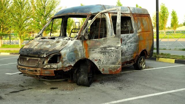 Burned van sits completely gutted on asphalt parking lot, showing charred metal frame and melted rubber tires. Broken windows and scorched panels reveal extensive damage to destroyed vehicle.