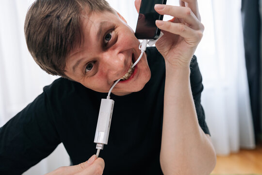 Man appearing stressed and dependent on technology, biting charging cable connected to smartphone, depicting addiction and desperate need for connection.