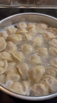 Close-up video of traditional meat dumplings pelmeni boiling in bubbling hot water inside a metal pot on an induction stove during cooking.