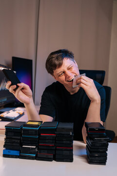 Playful content creator bites memory card, winking at camera. Man sitting at desk surrounded by stacks of external SSD hard drives, symbolizing data consumption, information overload or backup.