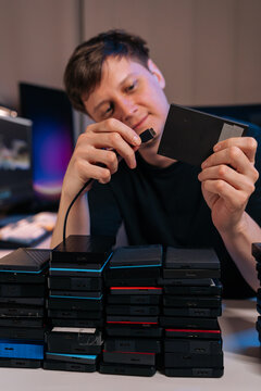 Focused technician connecting portable hard drive. Many external drives stacked on desk represent big data, information backup, storage management, or digital archiving. Soft lighting from screens.