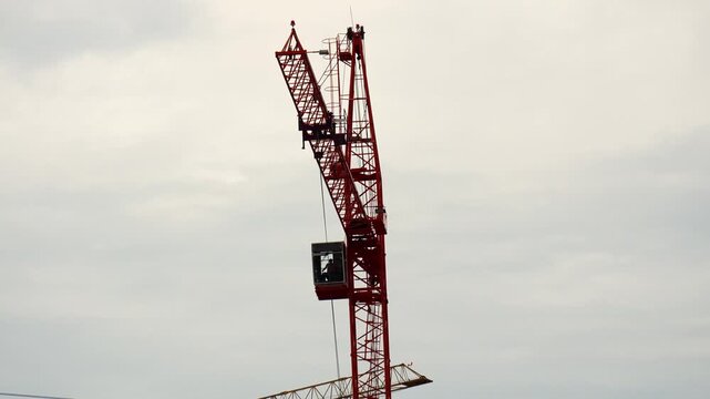 Industrial red luffing crane turns slowly across cloudy sky at modern building site. Steel construction equipment continues essential development operations with high engineering precision.