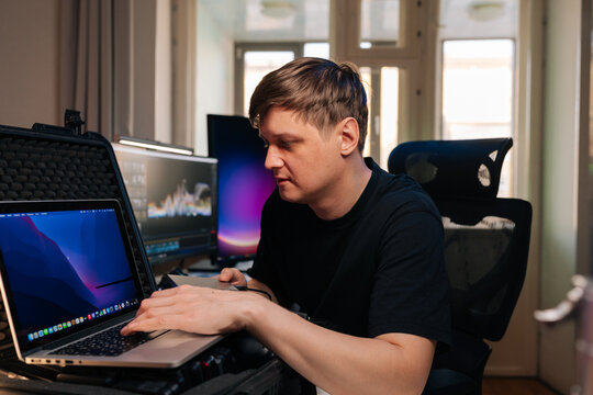 Focused man works on laptop inside protective case. Video editor or content creator in home studio with dual monitors. Professional freelancer concentrating on post production project.