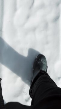 Vertical point-of-view video of feet in black warm rubber boots walking on deep white snow. Winter outdoor activity, walking in snowy field, cold weather travel.