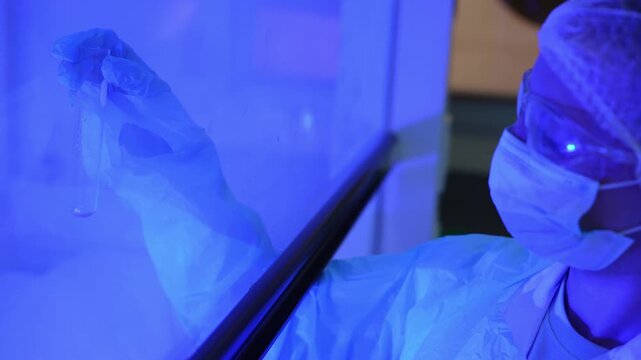 Scientist in mask and goggles holds up a test tube to inspect a small sample behind the sash of an ultraviolet biosafety cabinet, then turns her head, blue light close-up