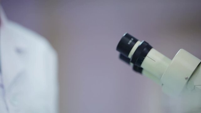Female scientist in white hair net and safety goggles looks into a microscope eyepiece, then leaves frame in a close-up rack of a quiet biotech laboratory analysis moment