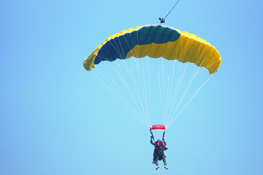 Back view of tandem skydiving team flying beneath yellow and blue parachute canopy. Symbolizes professional air adventure guidance, extreme sports recreation, and parachute flight control.